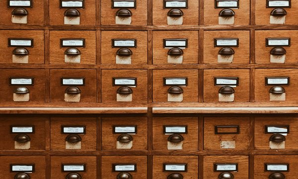 Holzkatalogschrank mit vielen Schubladen, jede versehen mit einem Metallschild und Griff, beschriftet mit polnischen Texten zur Kategorisierung, in einer Bibliotheksumgebung.