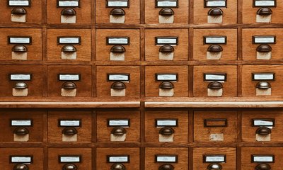 Holzkatalogschrank mit vielen Schubladen, jede versehen mit einem Metallschild und Griff, beschriftet mit polnischen Texten zur Kategorisierung, in einer Bibliotheksumgebung.