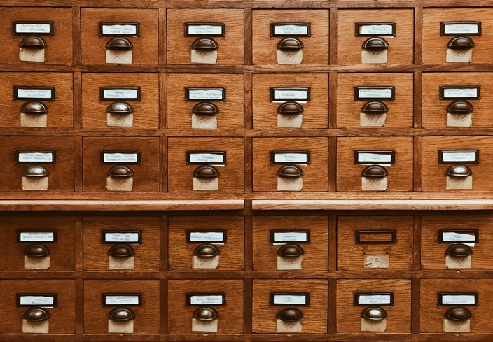 Holzkatalogschrank mit vielen Schubladen, jede versehen mit einem Metallschild und Griff, beschriftet mit polnischen Texten zur Kategorisierung, in einer Bibliotheksumgebung.