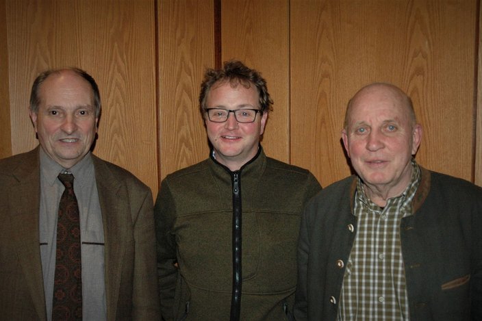 Drei Männer stehen nebeneinander vor einer Holzverkleidung. Sie lächeln in die Kamera, wobei der Mann in der Mitte eine Brille trägt und eine grüne Jacke anhat. Die beiden anderen tragen formelle Kleidung.